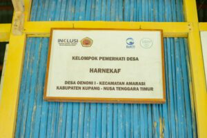 A framed sign reading “Kelompok Pemerhati Desa Harnekaf” hangs on the blue and yellow exterior wall of a building. The sign identifies the group as being based in Oenoni II Village, Amarasi District, Kupang Regency, East Nusa Tenggara, and displays the logos of the INKLUSI program and partner organizations.