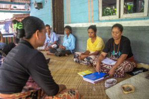 Wilhelmina, a woman wearing a black shirt seated on the right, holds and points to a blue scrapbook during a focus group discussion. In front of her, several women of different ages sit on a woven mat, including women wearing light blue and yellow clothing, while another participant is seen from behind in the foreground. 