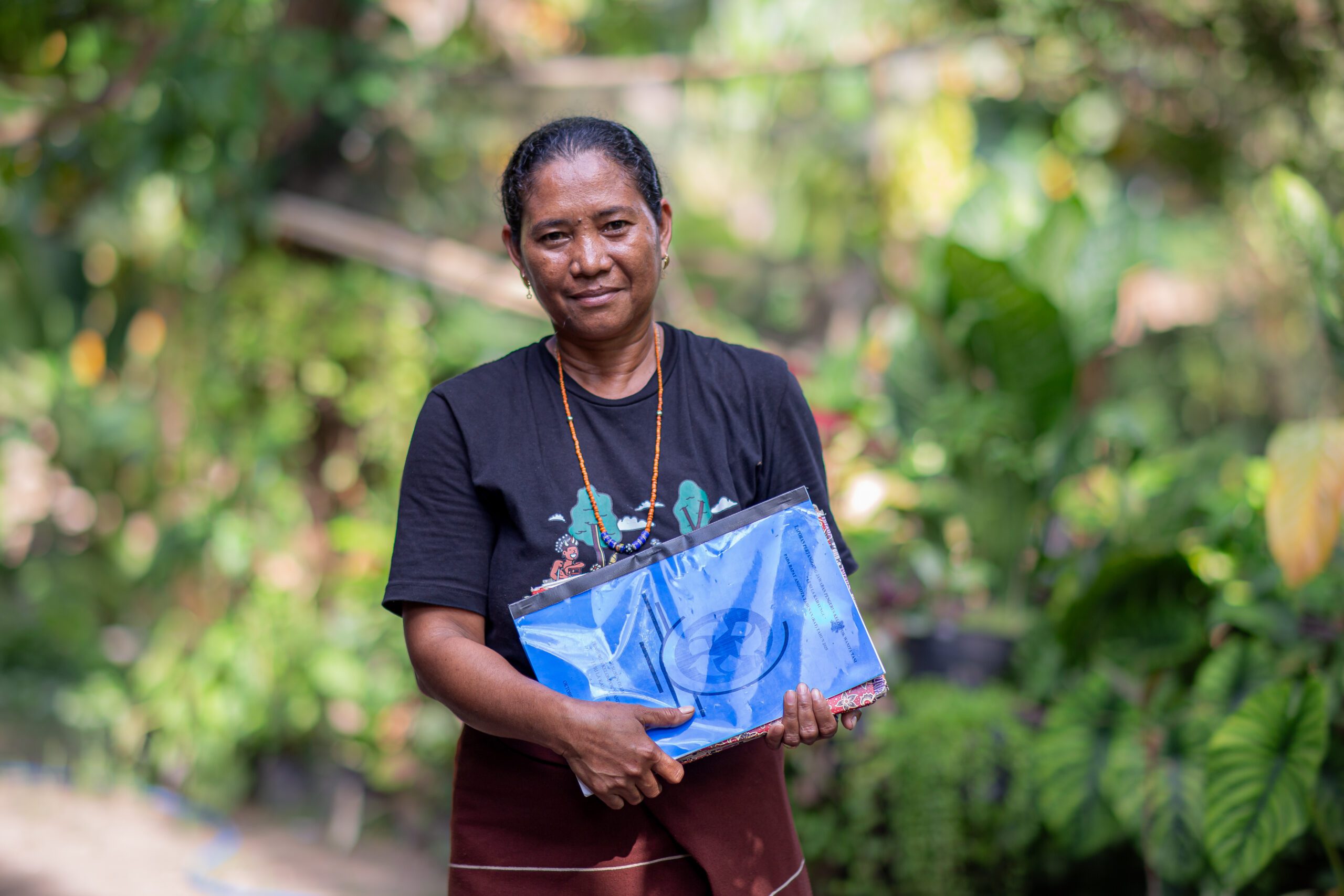 Wilhelmina, holding a scrapbook against the backdrop of a rural village, smiles broadly at the camera.