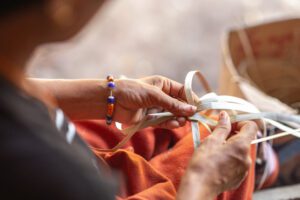 Close-up of two pairs of hands weaving long, pale strips of material together. One person wears a beaded bracelet, while the weaving rests against a red cloth in the background. 