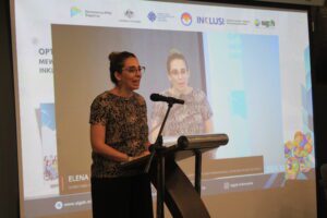Elena Martin Avila, First Secretary Kedutaan Besar Australia untuk Indonesia, berbicara tentang pentingnya ketenagakerjaan inklusif, di depan sebuah podium, dengan latar belakang siaran langsung kegiatan peluncuran ULD.
