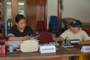 Ratna and Henny sit at a table discussing and reviewing documents. Ratna takes notes while holding a phone, while Henny, wearing a black cap, holds a document. A nameplate reading “BENDAHARA” and various documents and stationery are visible on the table.