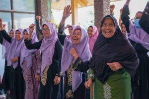 A group of women wearing purple and black clothing and headscarves raise their hands together during a lively community activity.