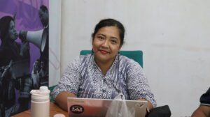 Safitri, Migrant CARE Project Manager, with black hair tied back, wearing a grey and white patterned blouse, sits in a green chair behind an open laptop during a meeting.