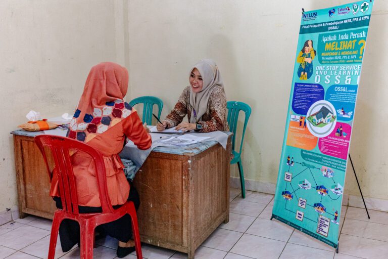 Seorang perempuan mengenakan jilbab dan pakaian berwarna oranye sedang berkonsultasi dengan seorang petugas perempuan di sebuah meja layanan. Petugas mencatat informasi pada dokumen di atas meja. Di sisi kanan terlihat poster informasi tentang layanan One Stop Service and Learning (OSS&L) untuk kesehatan seksual dan reproduksi, serta pencegahan dan penanganan kekerasan terhadap perempuan dan anak.